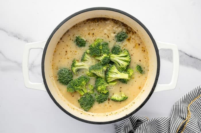 Overhead view of a white enameled Dutch oven filled with creamy soup base speckled with herbs. Fresh broccoli florets are scattered across the center of the pot, ready to simmer. The pot sits on a white marble countertop, with a black-and-white striped towel at the bottom right.