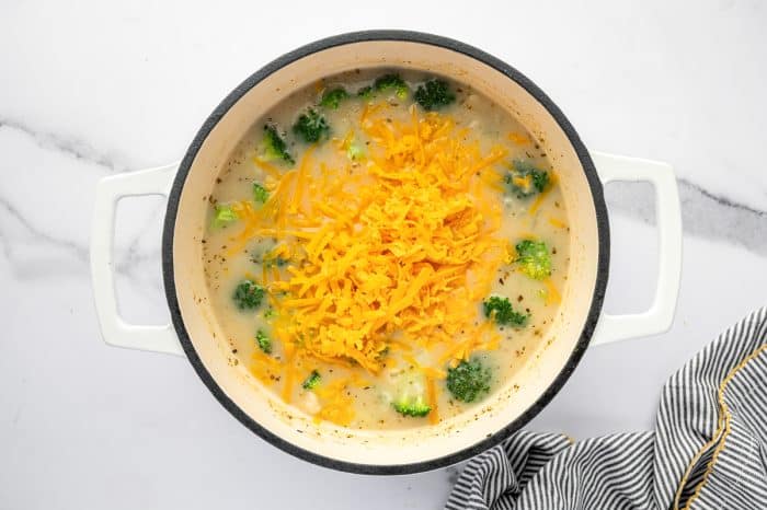 Overhead view of a white enameled Dutch oven filled with creamy broccoli potato soup. Broccoli florets and potato chunks are visible around the edges, and a large mound of shredded orange cheddar cheese sits in the center, ready to melt. A black-and-white striped kitchen towel appears at the bottom right on a white marble countertop.