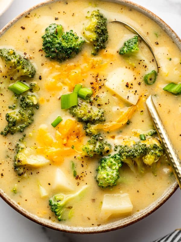 Extreme close-up of creamy broccoli potato soup in a gray bowl with a brown rim. Broccoli florets and potato chunks are visible beneath the surface, with melted cheddar pooled in the center and sliced green onions scattered on top. Cracked black pepper is sprinkled across the soup, and a metal spoon rests along the right side of the bowl.
