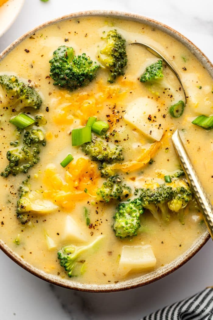 Extreme close-up of creamy broccoli potato soup in a gray bowl with a brown rim. Broccoli florets and potato chunks are visible beneath the surface, with melted cheddar pooled in the center and sliced green onions scattered on top. Cracked black pepper is sprinkled across the soup, and a metal spoon rests along the right side of the bowl.