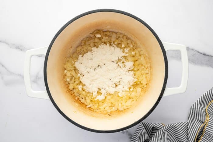 Overhead view of a white enameled Dutch oven with a dark rim and white handles on a white marble countertop. Softened diced onions fill the bottom of the pot, and a mound of all-purpose flour is sprinkled over the center to start the roux. A black-and-white striped kitchen towel is visible at the bottom right.