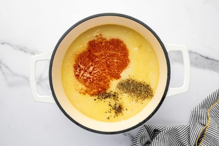 Overhead view of a white enameled Dutch oven on a white marble countertop filled with pale, creamy liquid. Seasonings are added in separate piles, including a large red mound of paprika, a small pile of dried green herbs (thyme), and scattered black pepper. A black-and-white striped towel sits at the bottom right edge of the frame.