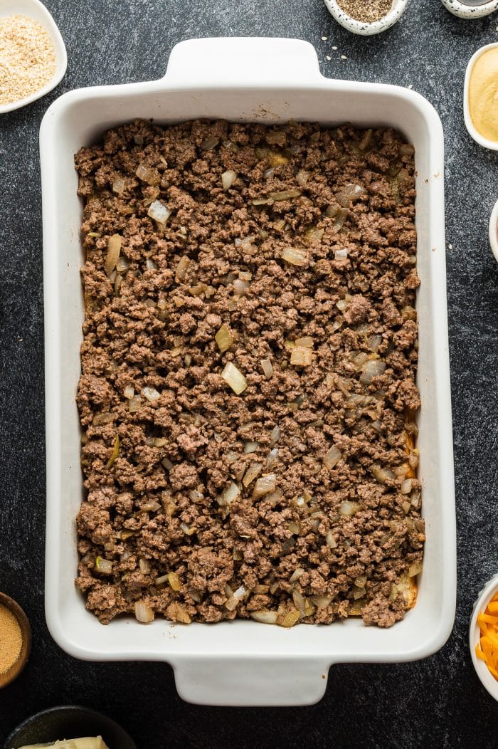 Overhead view of a white 9x13-inch baking dish filled with an even layer of browned ground beef and diced onions spread across the surface. The dish sits on a dark gray textured background with ingredient bowls partially visible at the edges, including sesame seeds near the top left and shredded cheddar near the right side.