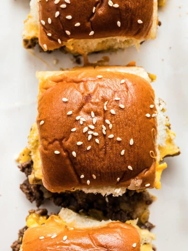 Overhead close-up of cheeseburger sliders arranged on a white platter, showing soft, shiny Hawaiian roll tops sprinkled with sesame seeds. Melted cheddar and browned ground beef are visible along the edges where the sliders have been cut, with a few cheese and beef bits scattered on the platter.