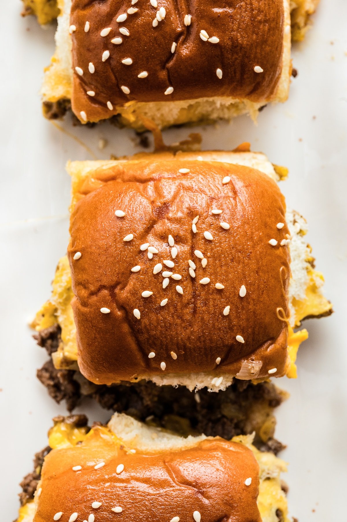 Overhead close-up of cheeseburger sliders arranged on a white platter, showing soft, shiny Hawaiian roll tops sprinkled with sesame seeds. Melted cheddar and browned ground beef are visible along the edges where the sliders have been cut, with a few cheese and beef bits scattered on the platter.