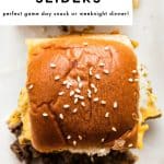 Vertical Pinterest-style image showing three cheeseburger sliders on a white platter, with the center slider in sharp focus. The shiny, golden-brown Hawaiian roll tops are sprinkled with sesame seeds, and melted cheddar and browned ground beef are visible around the edges. A large white text box overlay at the top reads โEASY CHEESEBURGER SLIDERSโ in bold black letters, with smaller black text underneath that says โperfect game day snack or weeknight dinner!โ and small black text near the bottom reads โthe forked spoon.โ