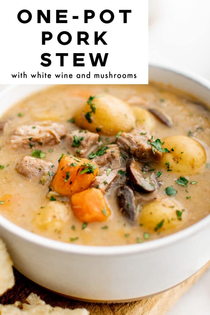 Pinterest pin showing a close-up of creamy one-pot pork stew in a white bowl, filled with baby potatoes, carrot chunks, sliced mushrooms, and tender pork pieces, sprinkled with chopped green herbs. A large white text box at the top left reads “ONE-POT PORK STEW” with the subtitle “with white wine and mushrooms.” Bread is partially visible along the left edge, and “theforkedspoon.com” appears as a small watermark at the bottom.