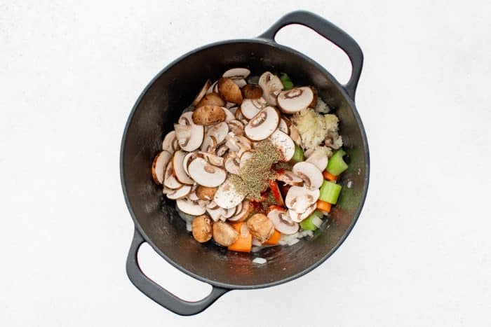 Overhead view of a black Dutch oven filled with sautéed vegetables topped with sliced mushrooms, a mound of minced garlic, dried thyme, and red paprika, all piled before stirring.