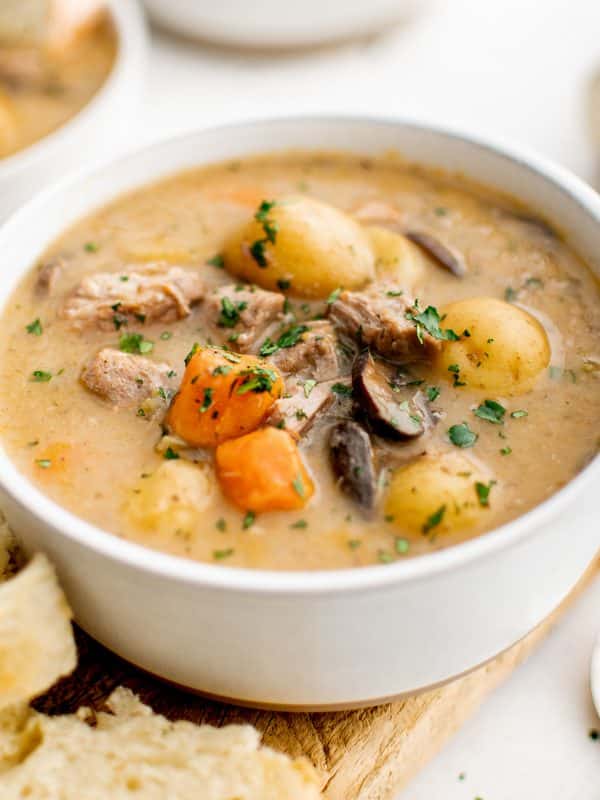 Close-up of a white bowl filled with creamy stovetop pork stew, showing tender pork pieces, baby potatoes, carrot chunks, and sliced mushrooms in a thick beige gravy, sprinkled with chopped green herbs, with torn bread in the foreground and a softly blurred bowl in the background.