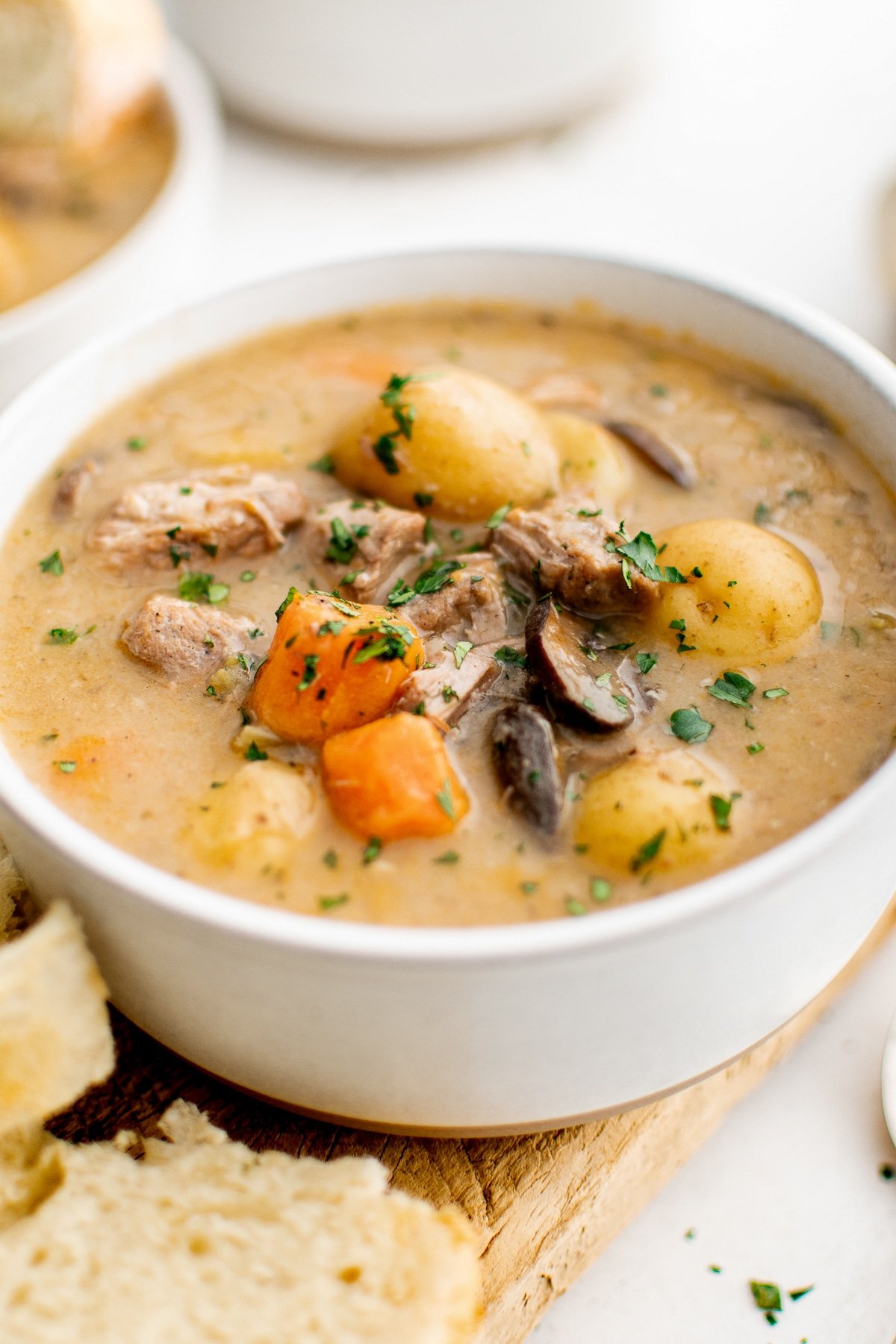 Close-up of a white bowl filled with creamy stovetop pork stew, showing tender pork pieces, baby potatoes, carrot chunks, and sliced mushrooms in a thick beige gravy, sprinkled with chopped green herbs, with torn bread in the foreground and a softly blurred bowl in the background.