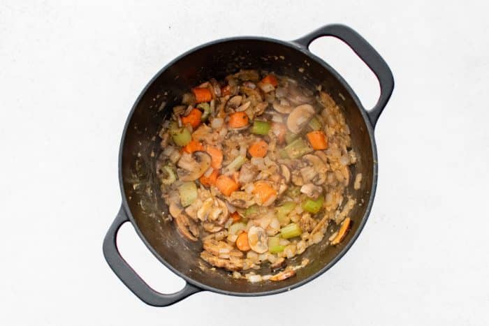 Overhead view of softened onions, carrots, celery, and mushrooms cooking in a black Dutch oven, lightly browned and steamy, with bits of sautéed vegetables stuck to the sides of the pot.