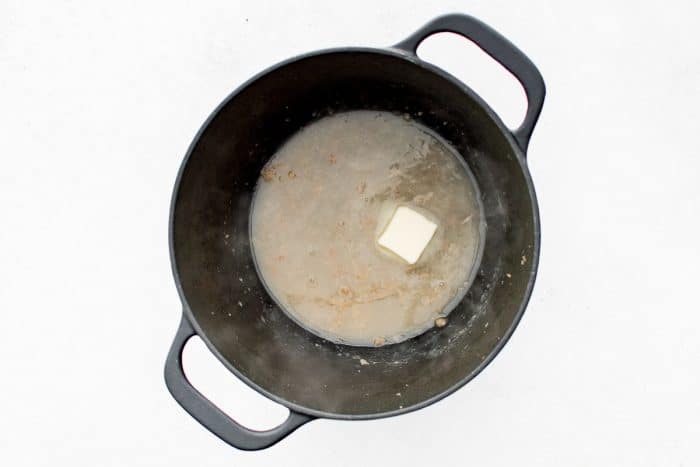 Overhead view of a black Dutch oven with pale deglazing liquid in the bottom and a single pat of butter melting in the center, on a white countertop with no text overlays.