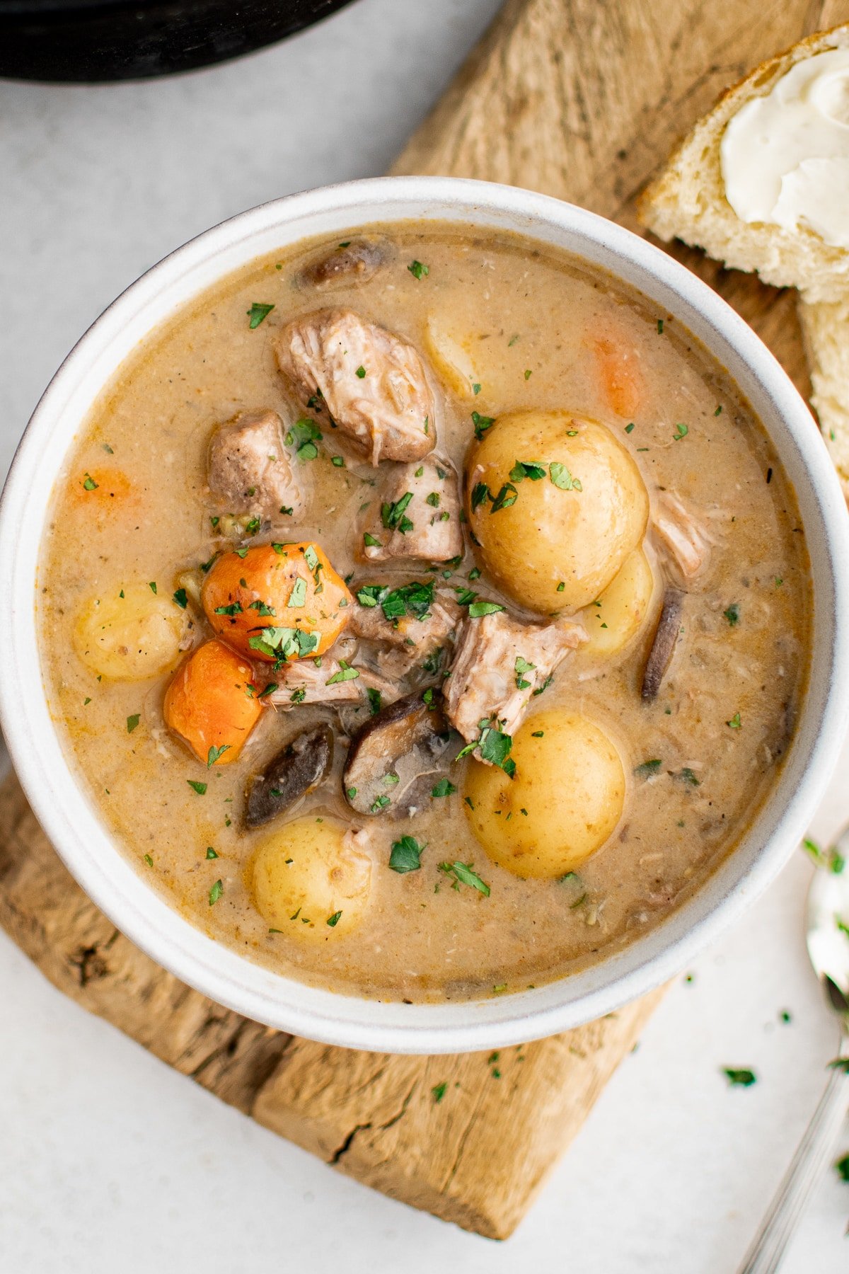Overhead view of a white bowl of creamy pork stew on a wooden board, filled with pork chunks, baby potatoes, carrots, and mushrooms in a thick beige gravy and sprinkled with chopped green herbs, with a piece of bread spread with butter visible at the top right and no text overlay.