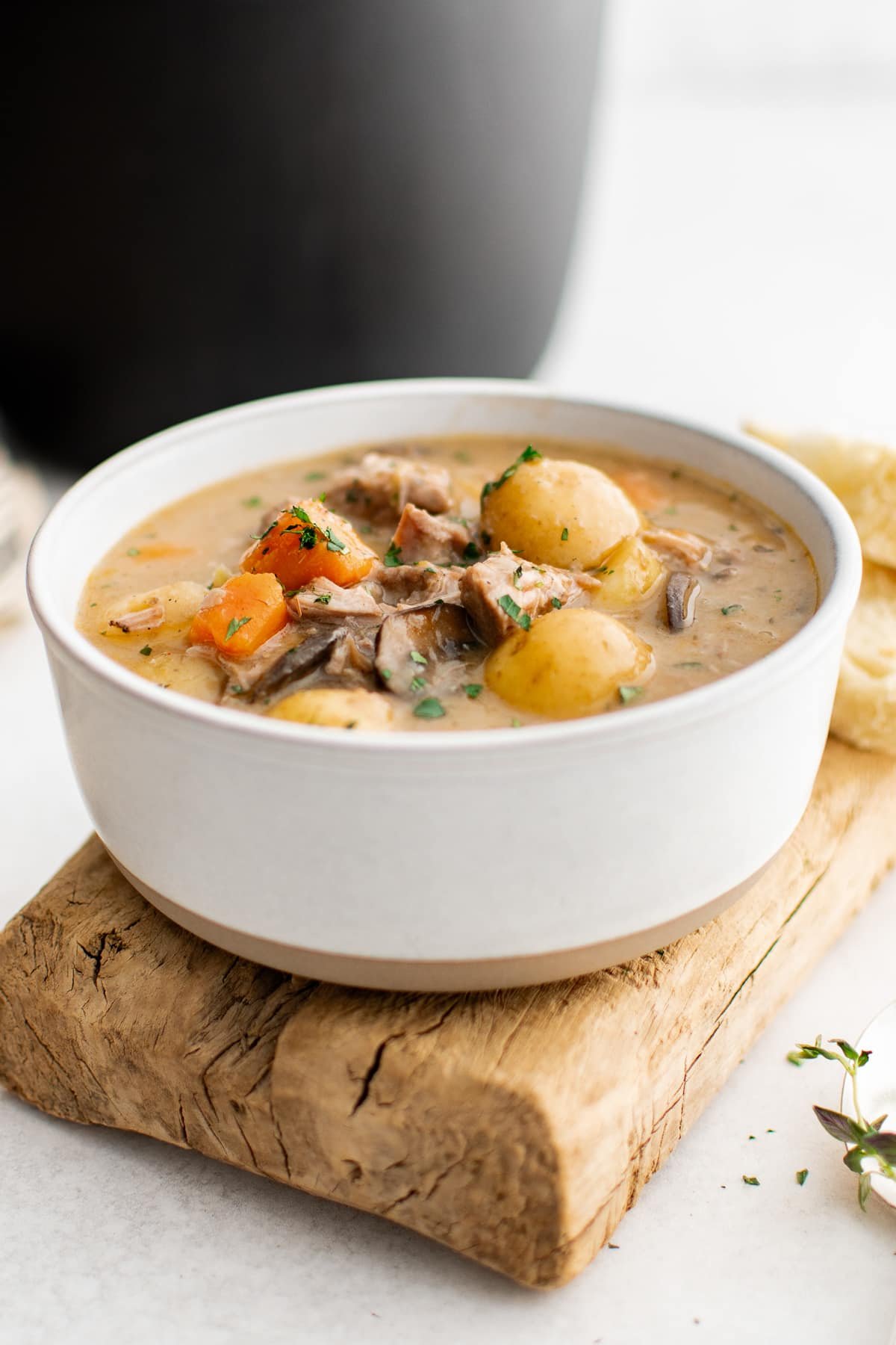 Side view of a white ceramic bowl filled with creamy pork stew on a rustic wooden board, with pork chunks, baby potatoes, carrots, and mushrooms visible in the thick gravy and topped with chopped green herbs, with a black pot softly blurred in the background.