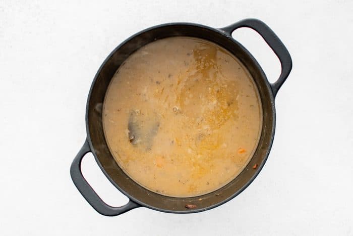 Overhead view of a black Dutch oven filled with a simmered, tan, gravy-like pork stew broth, lightly steaming with small bubbles at the surface and no garnishes or text overlays.