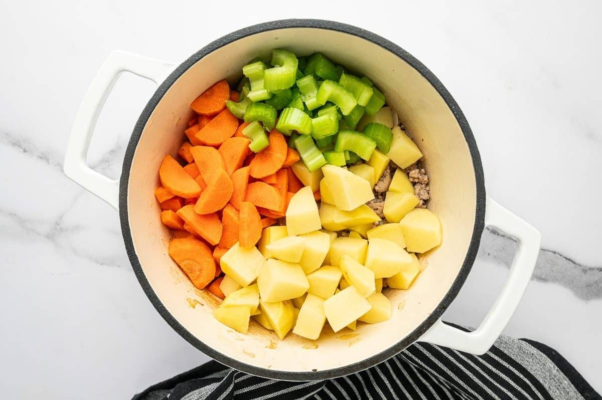 Overhead photo of sliced carrots, chopped celery, and peeled cubed potatoes added to browned ground turkey in a white enameled Dutch oven with a cream interior and black rim, on a white marble surface with a black-and-white striped towel underneath.