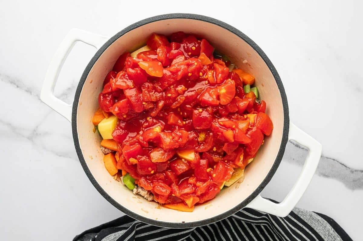 Overhead photo of canned diced tomatoes piled over carrots, potatoes, celery, and browned ground turkey in a white enameled Dutch oven with a cream interior and black rim, on a white marble background with a black striped towel.