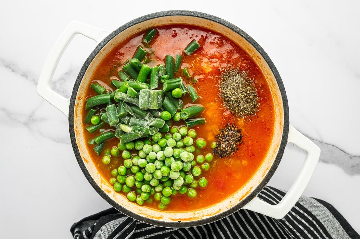 Overhead photo of red tomato broth in a white enameled Dutch oven with a cream interior and black rim, topped with frozen cut green beans and frozen peas, plus a mound of dried Italian seasoning and cracked black pepper on the surface; white marble background and black striped towel below the pot.