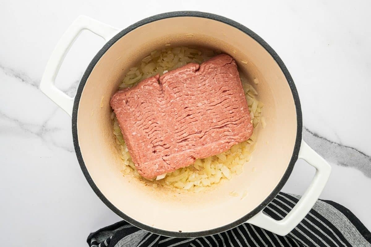 Overhead photo of a block of raw ground turkey placed on top of softened chopped onions and garlic in a white enameled Dutch oven with a cream interior and black rim on a white marble background with a black striped towel.