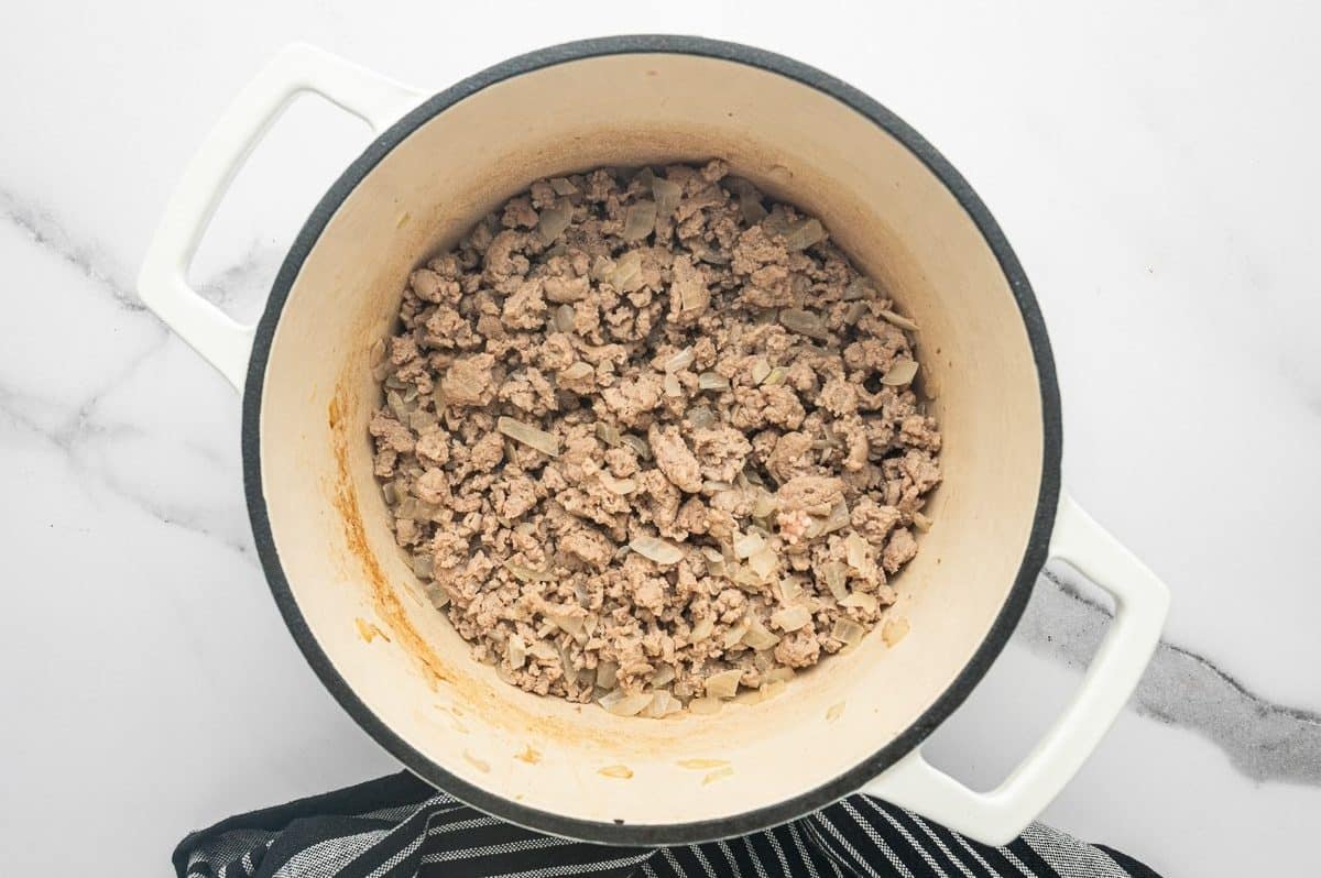 Overhead photo of browned crumbled ground turkey mixed with softened chopped onions inside a white enameled Dutch oven with a cream interior and black rim, with light browning on the sides of the pot, on a white marble surface and black striped towel.