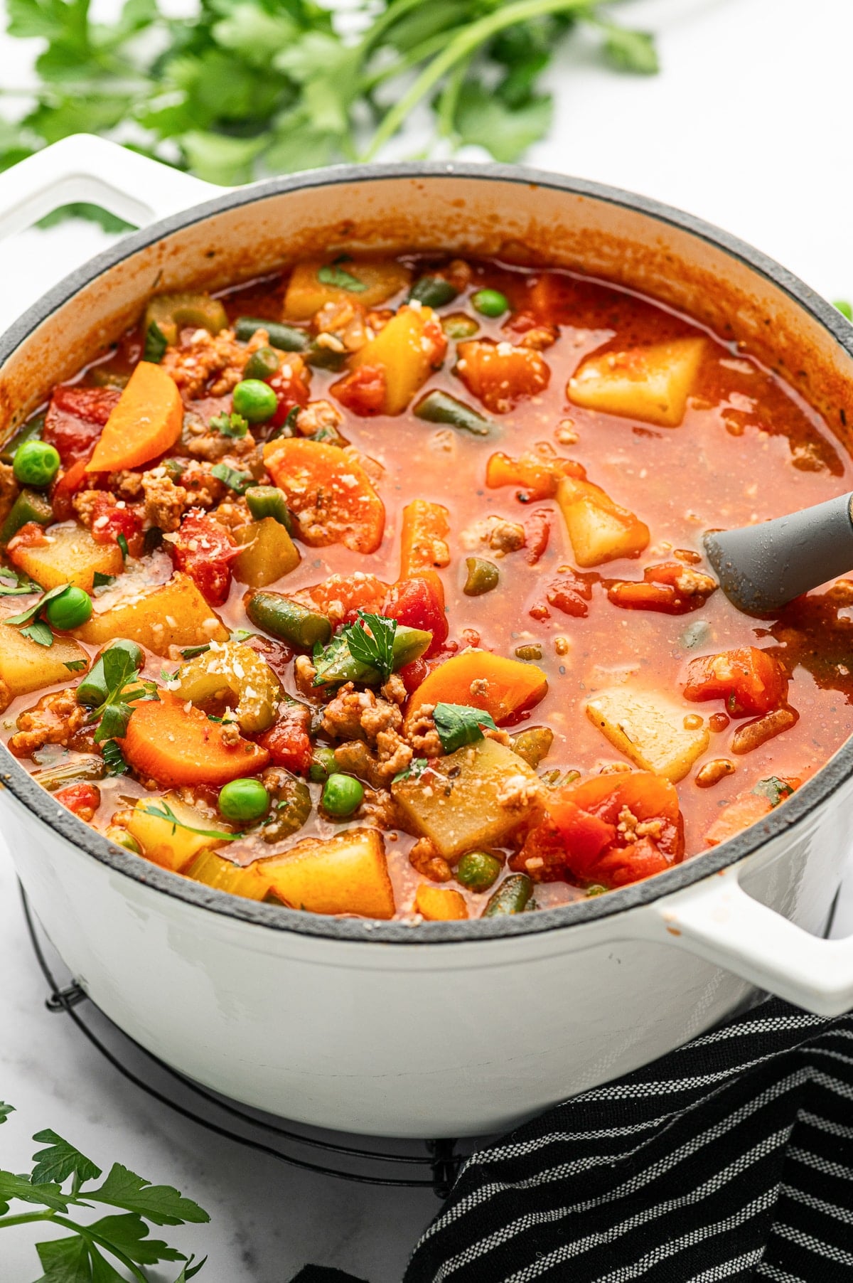 Close-up angled photo of ground turkey soup in a white enameled Dutch oven with a cream interior and black rim, showing a gray ladle resting in the red broth with visible potato chunks, sliced carrots, green beans, peas, and crumbled turkey; fresh parsley is blurred in the background and a black striped towel sits beside the pot.