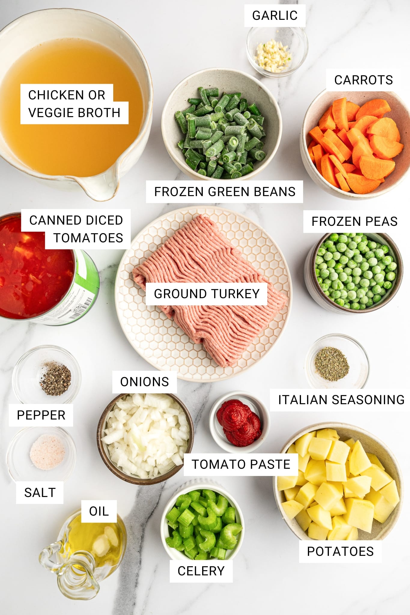 Overhead ingredients photo on a white marble surface with white text labels on each item: a glass measuring cup filled with golden chicken or veggie broth labeled “CHICKEN OR VEGGIE BROTH,” a small glass bowl of minced garlic labeled “GARLIC,” a bowl of sliced carrots labeled “CARROTS,” a bowl of frozen cut green beans labeled “FROZEN GREEN BEANS,” a bowl of frozen peas labeled “FROZEN PEAS,” a hexagon-pattern plate with raw ground turkey labeled “GROUND TURKEY,” a bowl of chopped onions labeled “ONIONS,” a small dish of tomato paste labeled “TOMATO PASTE,” a small dish of dried herbs labeled “ITALIAN SEASONING,” a bowl of cubed potatoes labeled “POTATOES,” a bowl of sliced celery labeled “CELERY,” a small glass dish of black pepper labeled “PEPPER,” a small glass dish of pink salt labeled “SALT,” a small glass bottle of oil labeled “OIL,” and an open can of diced tomatoes labeled “CANNED DICED TOMATOES.”