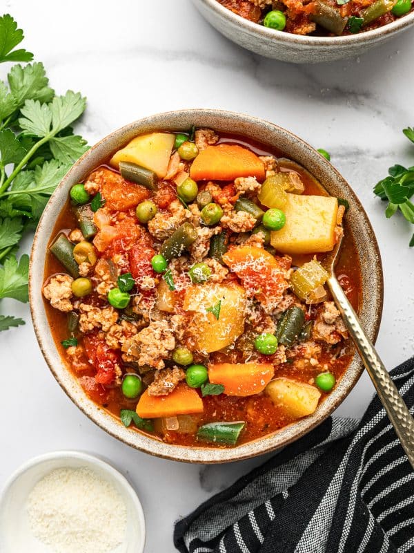 Overhead photo of a gray ceramic bowl filled with ground turkey soup, showing potatoes, carrots, green beans, peas, and turkey in a red broth, topped with chopped parsley and grated Parmesan; a gold spoon rests in the bowl, with a small white bowl of Parmesan, parsley sprigs, and a black striped towel on a white marble background.