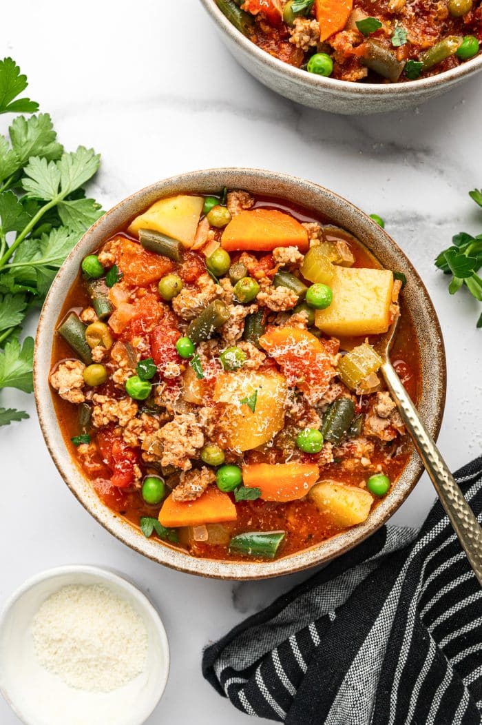 Overhead photo of a gray ceramic bowl filled with ground turkey soup, showing potatoes, carrots, green beans, peas, and turkey in a red broth, topped with chopped parsley and grated Parmesan; a gold spoon rests in the bowl, with a small white bowl of Parmesan, parsley sprigs, and a black striped towel on a white marble background.