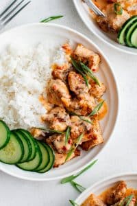 Overhead photo of a white bowl with fluffy white rice, Bang Bang salmon bites drizzled with creamy sauce, sliced green onions, and sliced cucumber rounds on the side, set on a light gray surface