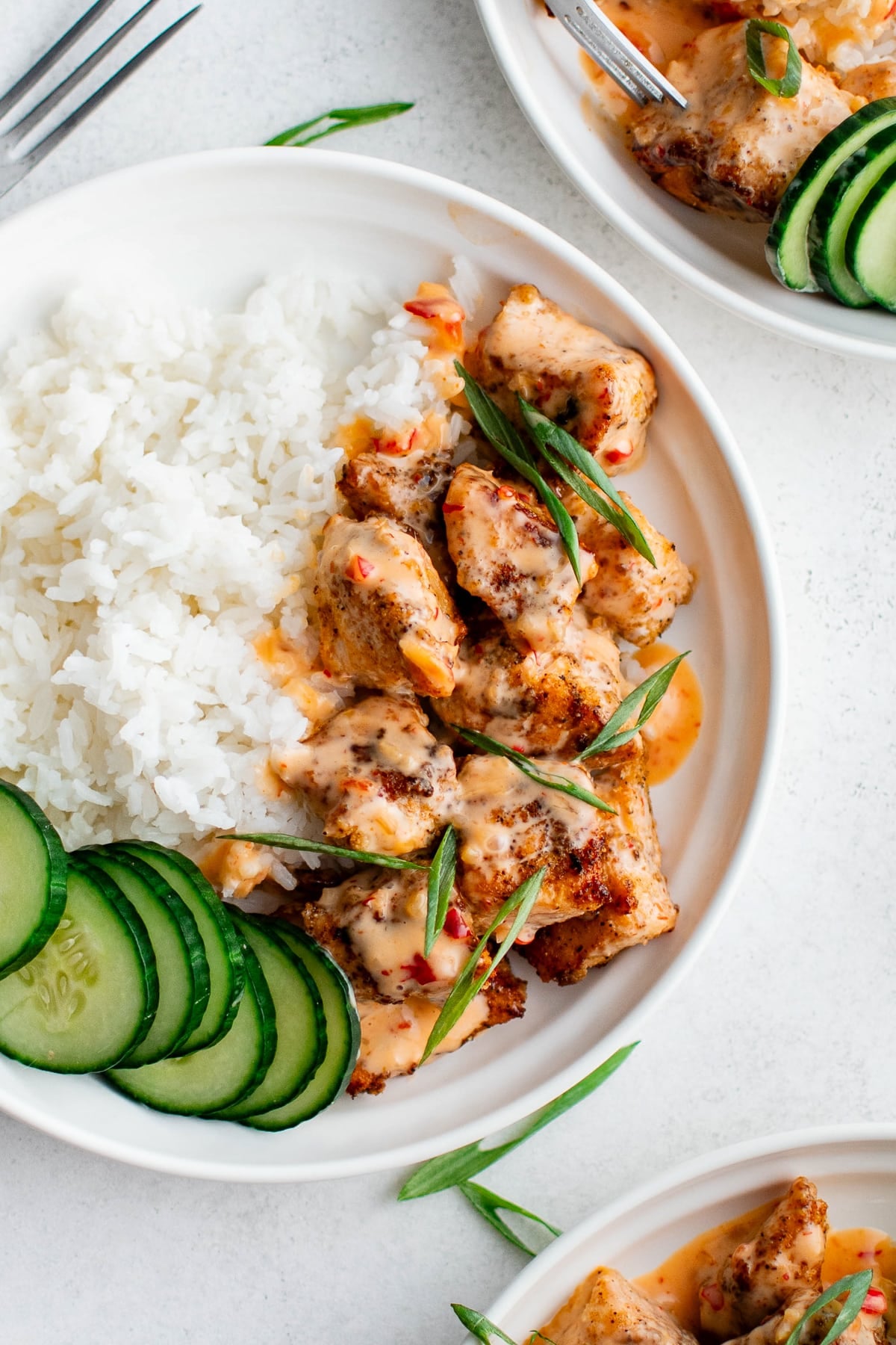 Overhead photo of a white bowl with fluffy white rice, Bang Bang salmon bites drizzled with creamy sauce, sliced green onions, and sliced cucumber rounds on the side, set on a light gray surface