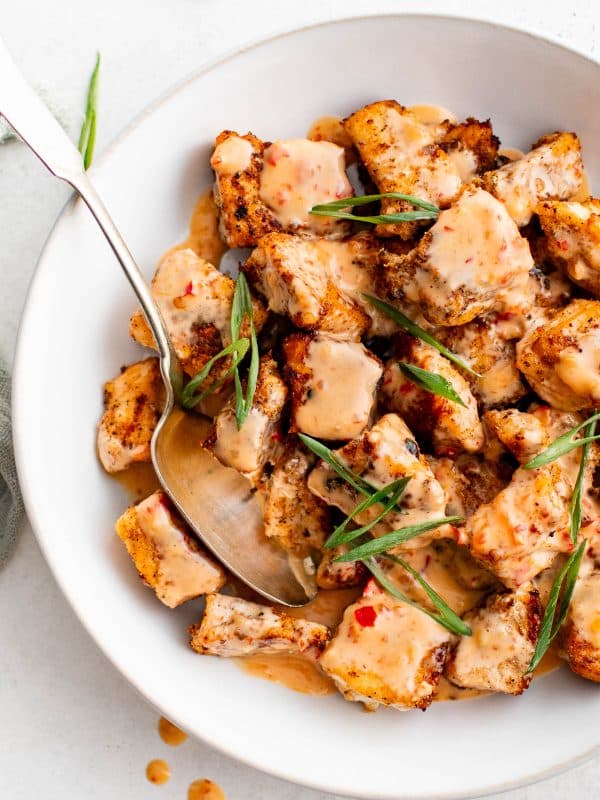 Close overhead photo of crispy salmon bites in a white bowl drizzled with creamy orange Bang Bang sauce and topped with sliced green onions, with a metal spoon resting in the bowl on a light background