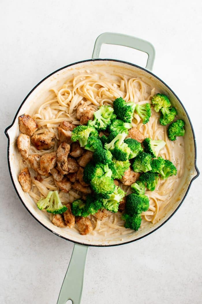 Overhead image of creamy fettuccine in a large white skillet with pale green handles topped with cooked bite-sized chicken pieces and bright green broccoli florets before tossing.