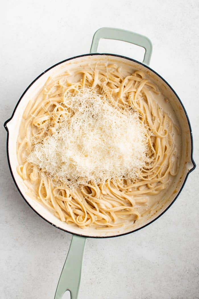 Overhead image of creamy fettuccine in a large white skillet with pale green handles topped with a mound of finely grated Parmesan cheese. The sauce is pale and creamy, coating the noodles evenly. The skillet sits on a light gray surface.