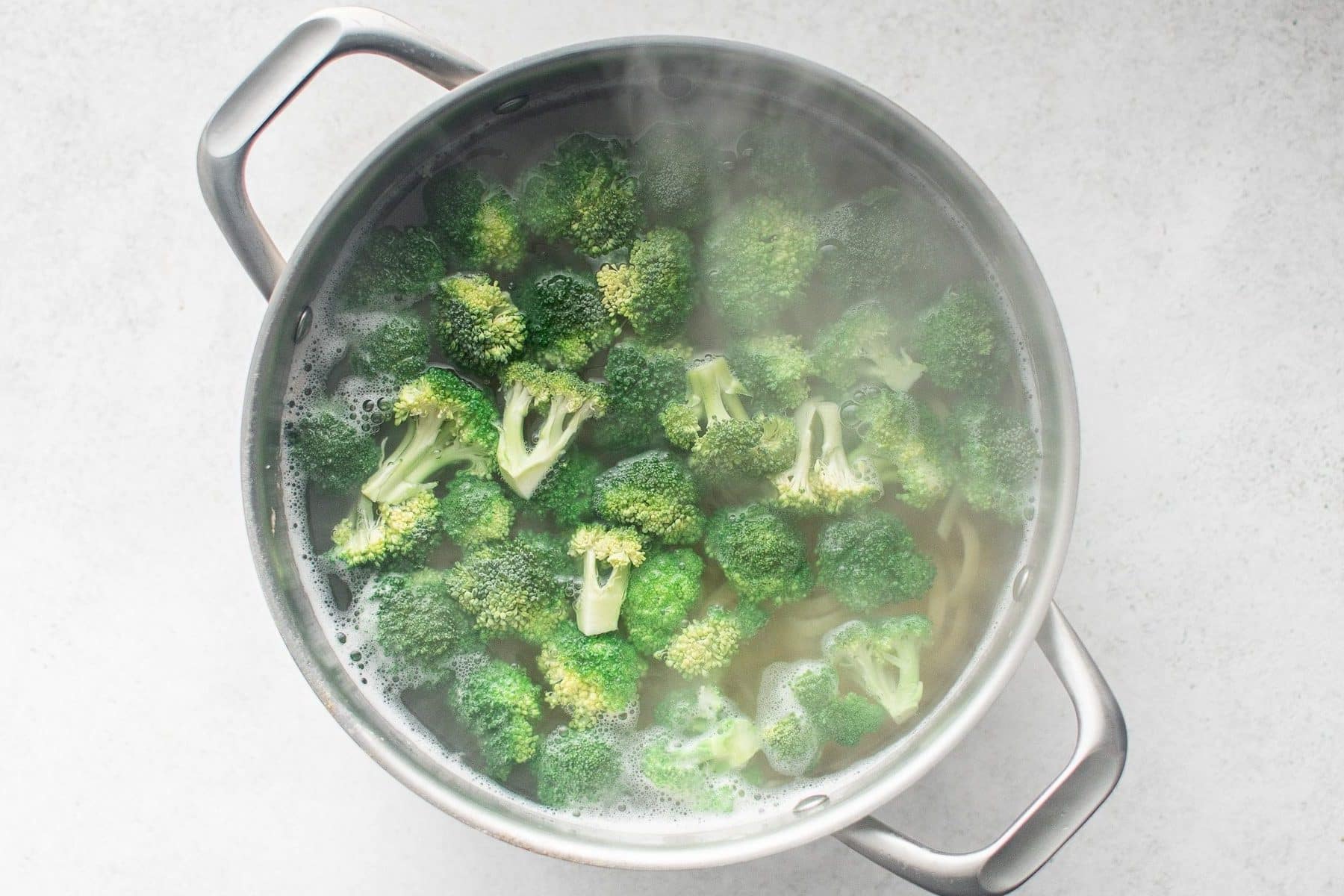 Overhead image of bright green broccoli florets blanching in a stainless steel pot of hot water with pasta underneath. Steam rises from the pot, and the broccoli is vibrant green and just tender. The pot sits on a light gray surface.