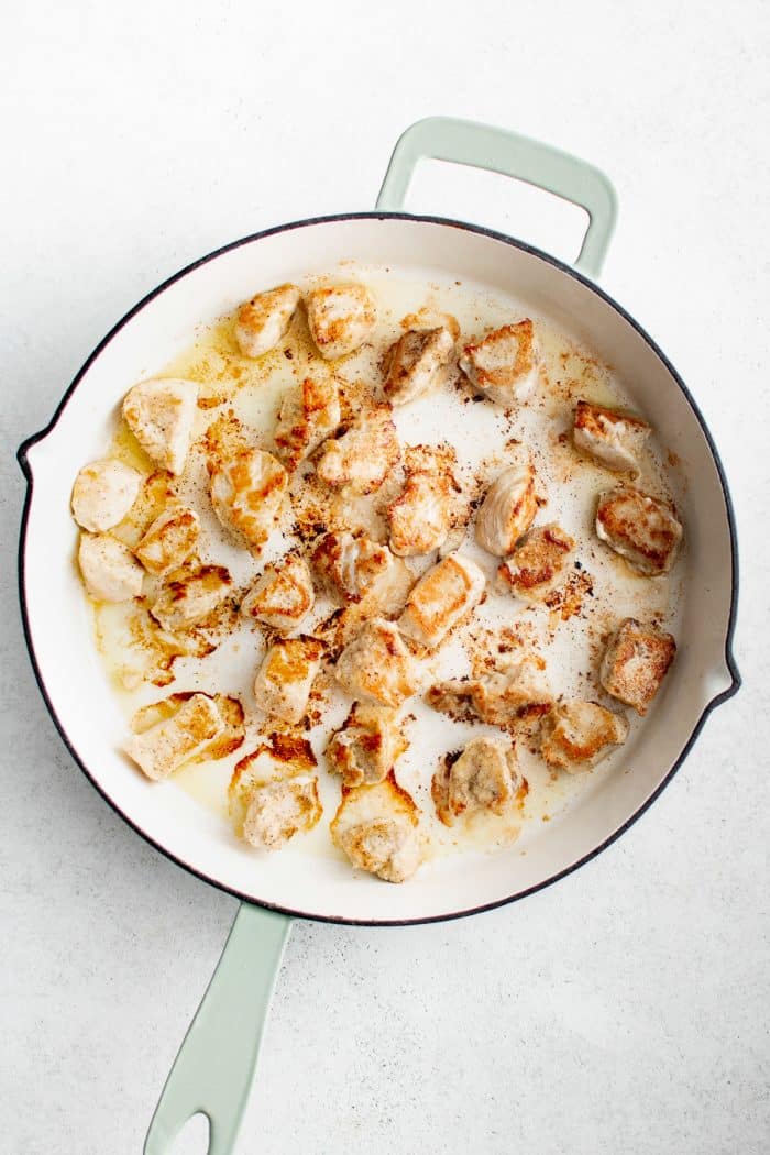 Overhead image of browned bite-sized chicken pieces cooking in a large white skillet with pale green handles. The chicken is golden brown in spots with light browning and cooking juices visible in the pan. The skillet sits on a light gray surface with no garnishes or text overlays.