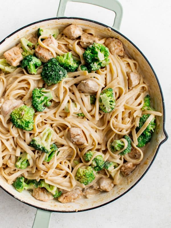 Overhead image of finished chicken broccoli Alfredo in a large white skillet with pale green handles. Creamy fettuccine is tossed with bite-sized chicken and bright green broccoli florets, all coated in a light Alfredo sauce.