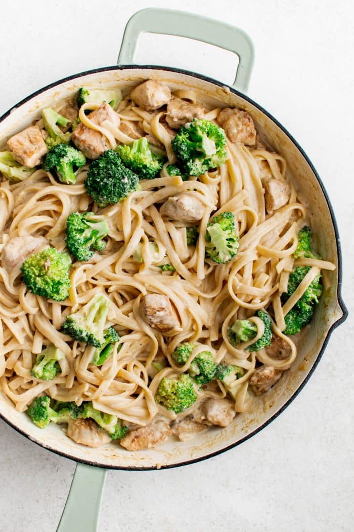 Overhead image of finished chicken broccoli Alfredo in a large white skillet with pale green handles. Creamy fettuccine is tossed with bite-sized chicken and bright green broccoli florets, all coated in a light Alfredo sauce.