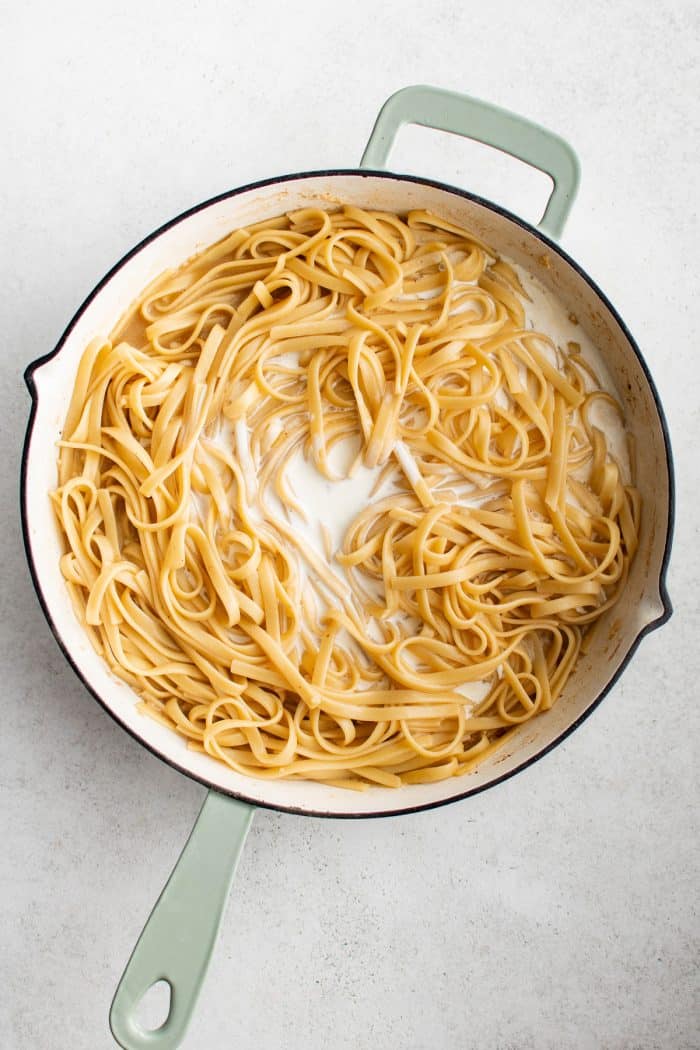 Overhead image of cooked fettuccine in a large white skillet with pale green handles, with heavy cream being added to the pan. The pasta sits in a shallow sauce base with cream pooling between the noodles. The skillet is on a light gray surface.