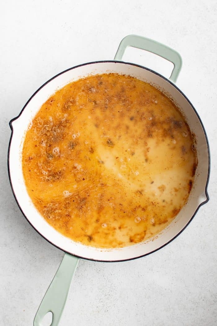 Overhead image of a light orange Alfredo-style sauce base simmering in a large white skillet with pale green handles. The sauce is made from pasta water, butter, garlic, with browned bits visible throughout the pan. The skillet is on a light gray surface,