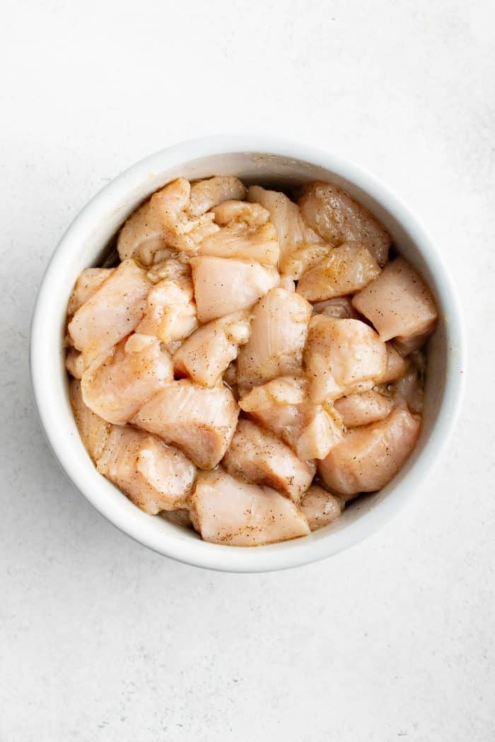 Overhead image of raw bite-sized chicken pieces seasoned with spices in a small white bowl on a light gray surface. The chicken is pale pink with visible black pepper and seasoning throughout.