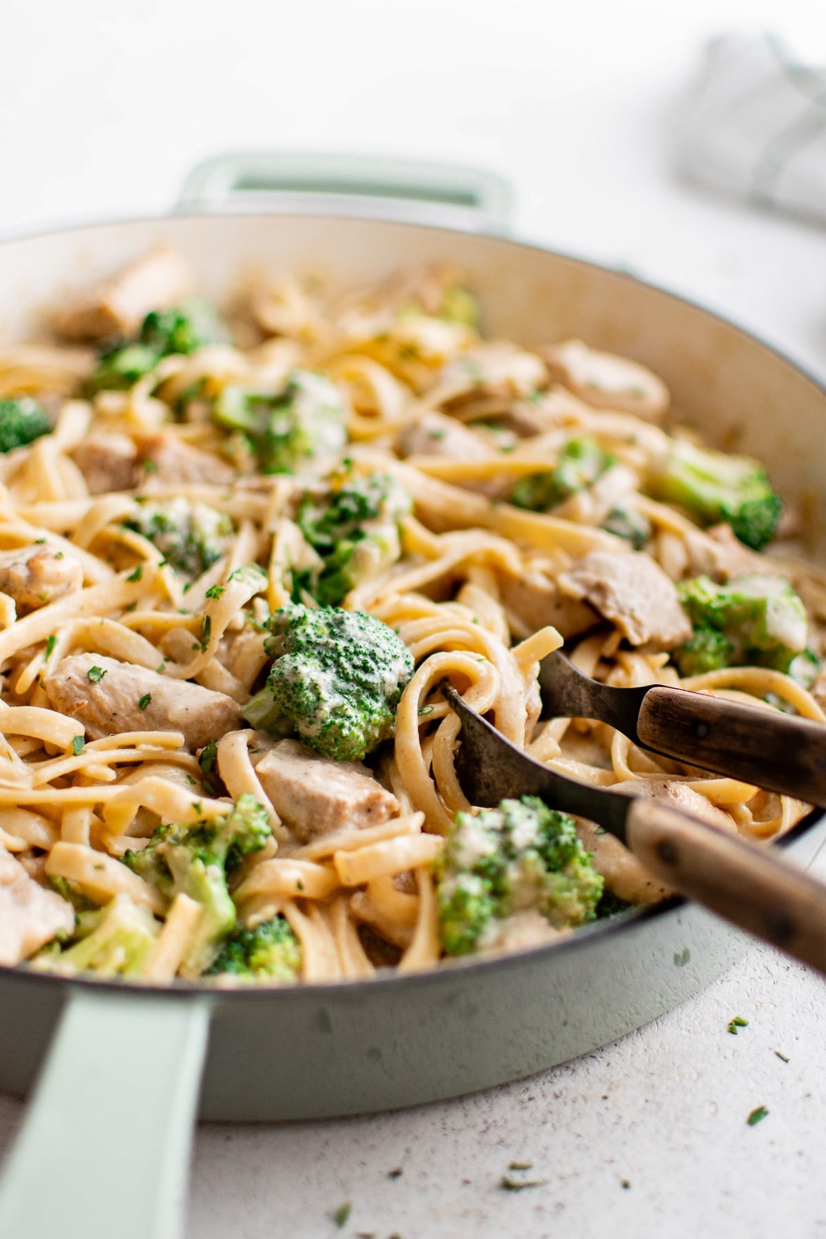Close-up image of chicken broccoli Alfredo served in a pale green-handled skillet with wooden pasta tongs resting in the pan. The creamy fettuccine is mixed with bite-sized chicken and broccoli florets and lightly sprinkled with chopped green herbs. The background is softly blurred, and there are no text overlays.