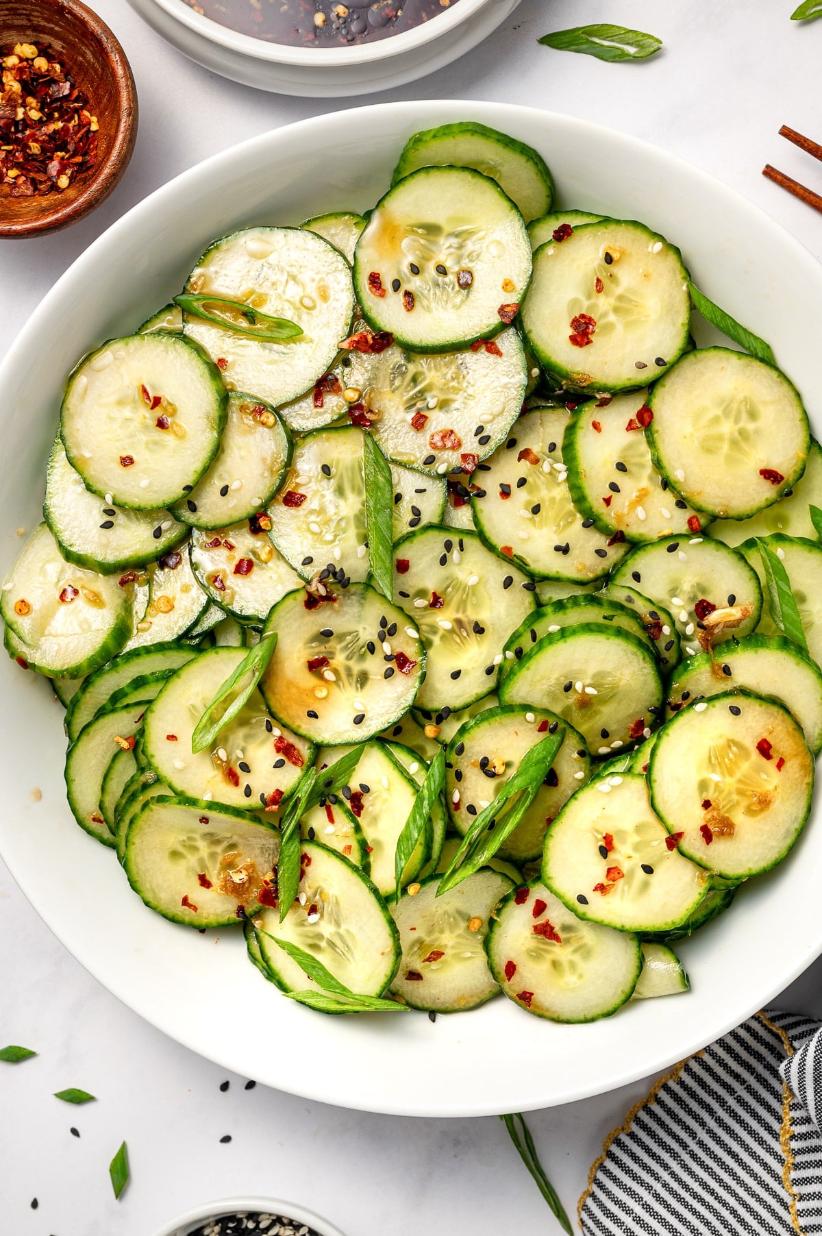 Close overhead image of Asian cucumber salad in a large white oval bowl, showing thin cucumber slices coated in dressing and topped with sliced green onions, black and white sesame seeds, and red pepper flakes, with partial bowls of dressing and red pepper flakes visible at the top and a gray and white striped towel with gold trim at the bottom edge.