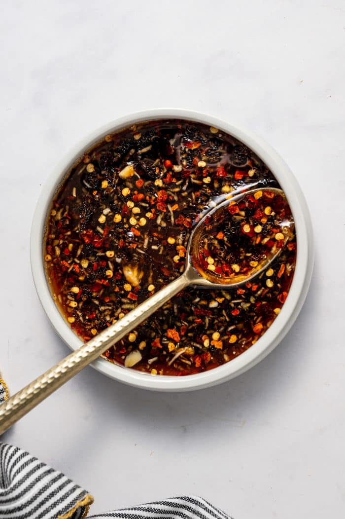 Overhead image of a small white bowl filled with mixed Asian cucumber salad dressing, with visible minced garlic, grated ginger, red pepper flakes, and dark soy-based liquid, with a gold spoon resting inside the bowl on a white marble surface and a gray and white striped towel with gold trim in the lower left corner.