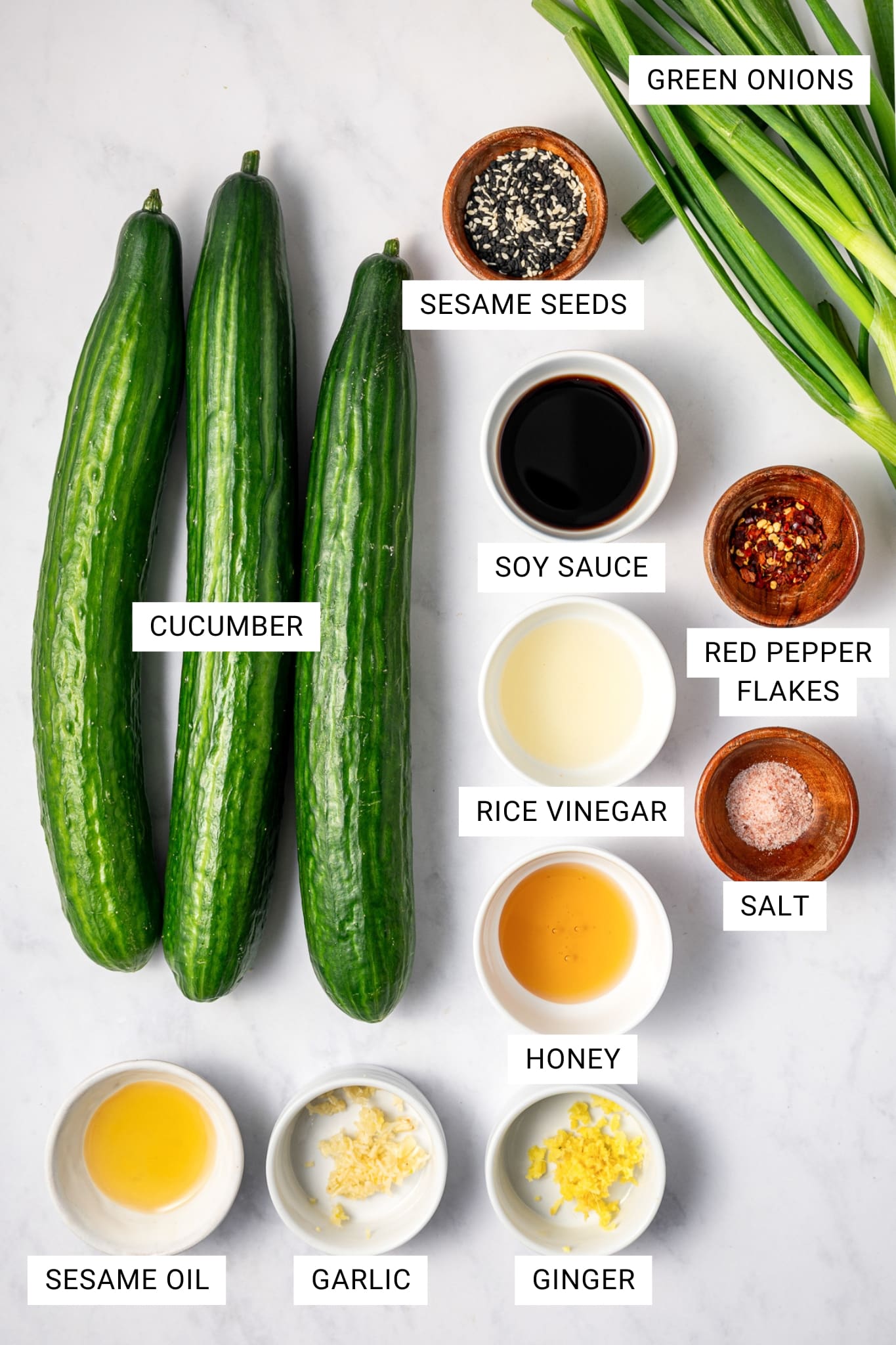 Overhead image of the ingredients for Asian cucumber salad arranged on a white marble surface with text overlays labeling each ingredient: cucumber, sesame seeds, soy sauce, red pepper flakes, rice vinegar, salt, honey, sesame oil, garlic, ginger, and green onions. Three whole cucumbers are arranged on the left, with small bowls of the remaining ingredients placed around them.