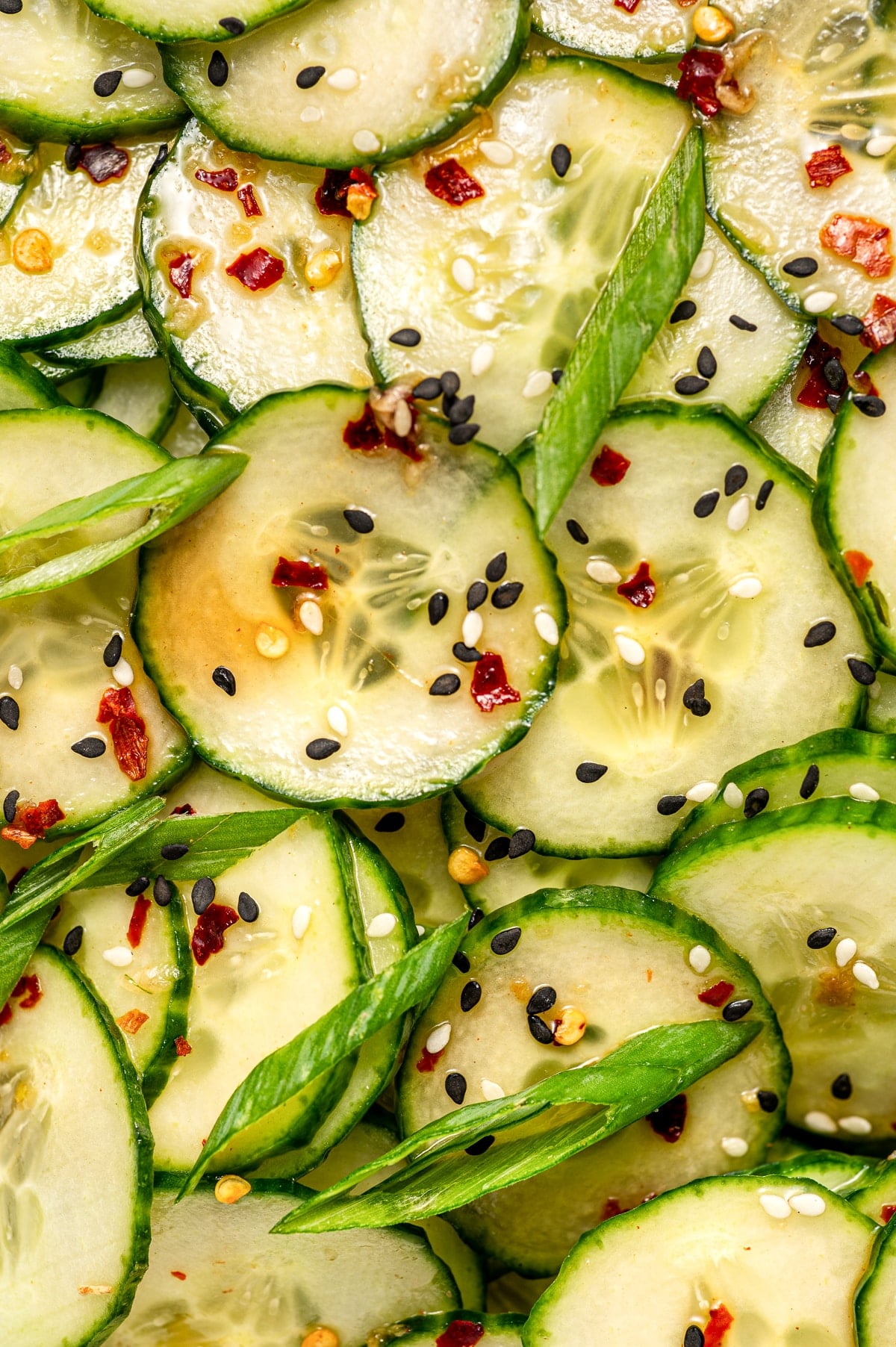 Extreme close-up image of Asian cucumber salad showing thin cucumber slices coated in dressing and topped with black and white sesame seeds, sliced green onions, and red pepper flakes.