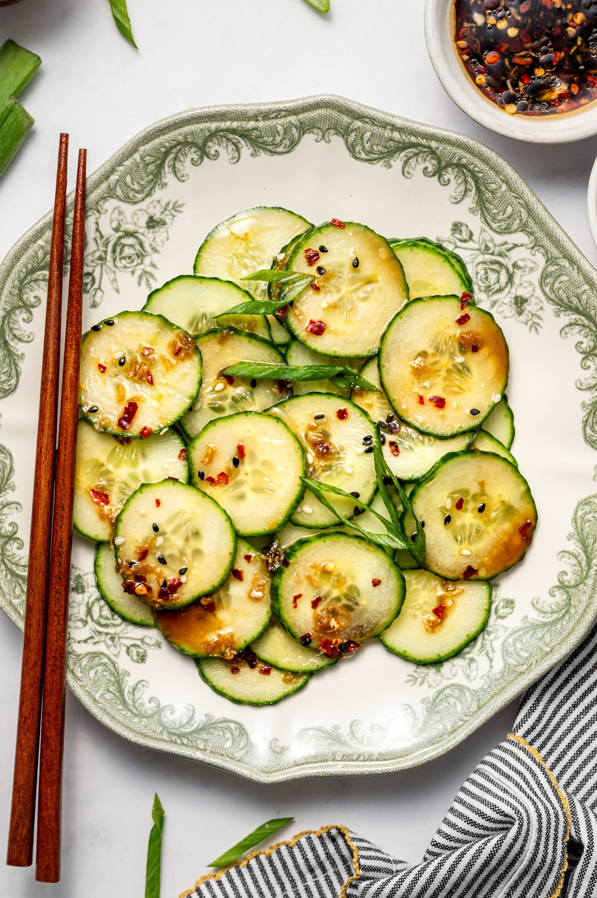 Overhead image of a portion of Asian cucumber salad served on a white plate with green floral trim, garnished with green onions, sesame seeds, and red pepper flakes, with wooden chopsticks on the left, small bowls of dressing nearby, scattered green onion slices, and a gray and white striped towel with gold trim on a white marble surface.