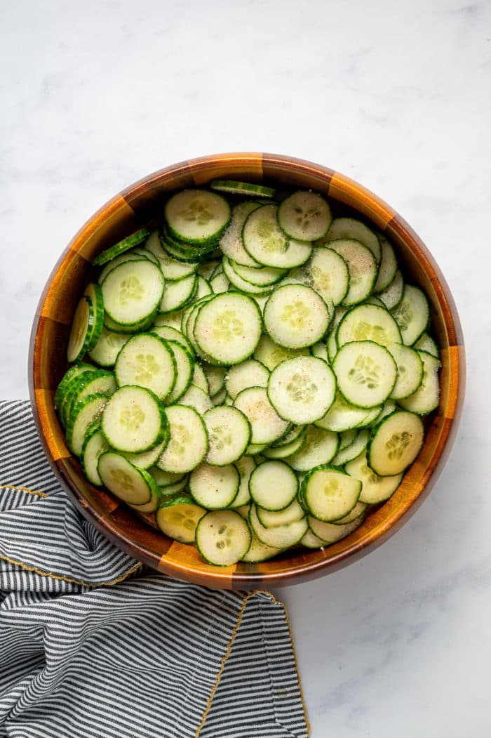 Overhead image of thinly sliced cucumbers sprinkled with salt in a round brown wooden bowl on a white marble surface, with a gray and white striped kitchen towel with gold trim placed to the right.