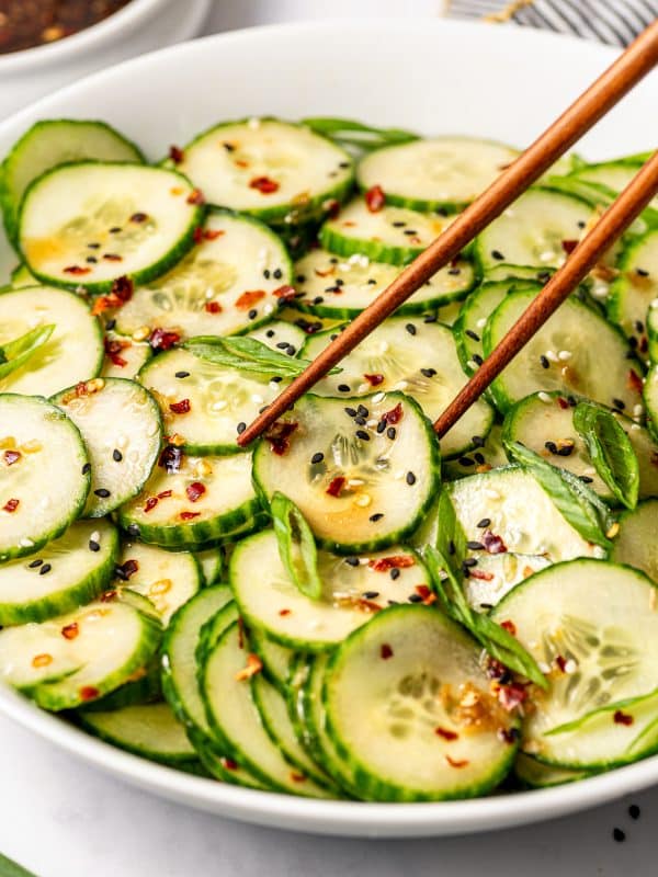 Angled close-up image of Asian cucumber salad in a large white bowl with wooden chopsticks lifting or reaching toward the cucumber slices, garnished with black and white sesame seeds, green onions, and red pepper flakes, with a bowl of dressing blurred in the background.