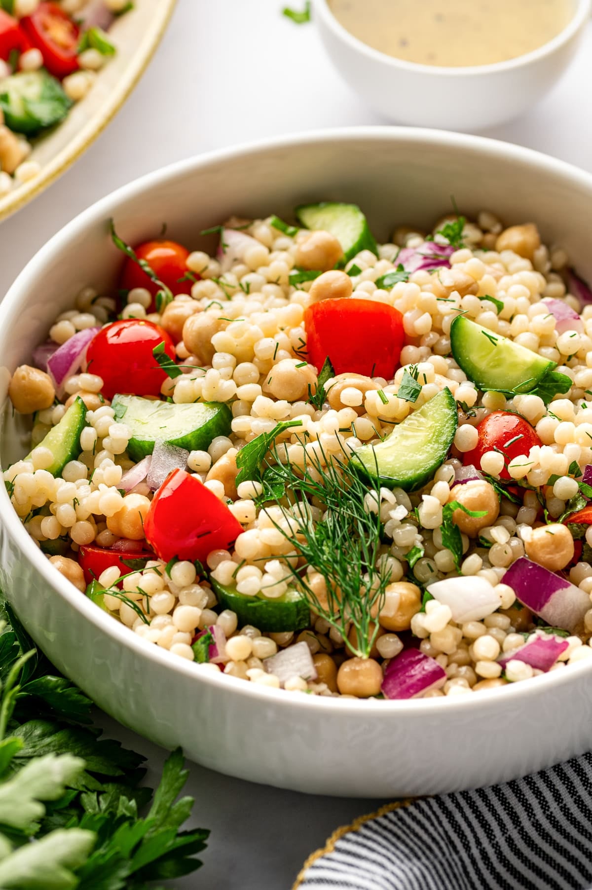 Close-up image of pearl couscous salad in a white bowl with visible chickpeas, halved cherry tomatoes, cucumber slices, diced red onion, chopped parsley, and fresh dill garnish on top. A small white bowl of dressing and part of a serving platter appear blurred in the background.