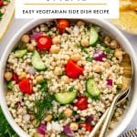 Vertical Pinterest image for couscous salad showing a white bowl filled with pearl couscous salad on a white background. The salad includes chickpeas, halved cherry tomatoes, chopped cucumber, diced red onion, parsley, and dill, with gold serving spoons resting in the bowl. A large white text box overlays the top of the image with black text that reads โCOUSCOUS SALADโ and smaller text below that reads โEASY VEGETARIAN SIDE DISH RECIPE,โ separated by a thin yellow line. Around the bowl are lemon wedges, a small white bowl of dressing, fresh herbs, part of a serving platter, and a gray and white striped towel with yellow trim. Theforkedspoon.com appears at the bottom.